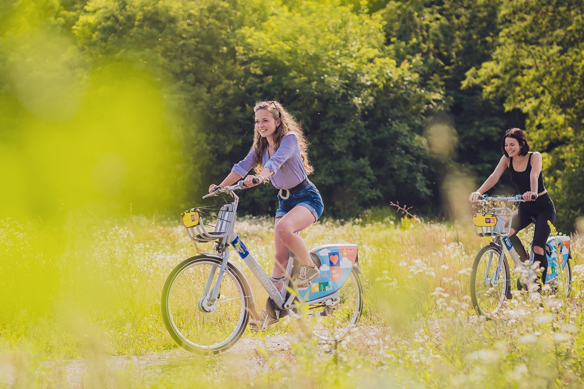 Dvě dívky jedoucí na sdílených kolech Nextbike loukou plnou květin
